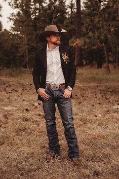 a man wearing a cowboy hat and jeans standing in the grass with his hands in his pockets