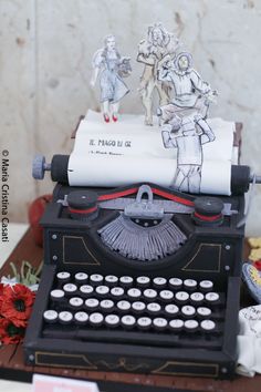 an old fashioned typewriter with paper cutouts on it's side and two figurines standing next to it