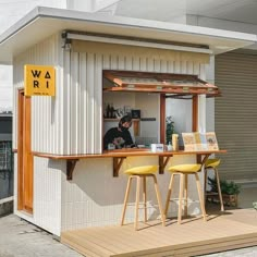 a man sitting at a table in front of a small white building with yellow stools