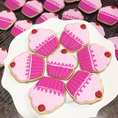 pink decorated heart shaped cookies on a white plate next to cooling rack with hearts and cupcakes