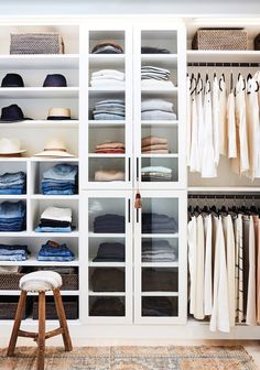 a white closet filled with lots of clothes and hats on top of glass shelves next to a stool