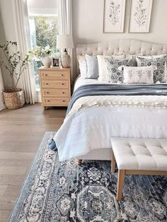 a white bed with blue and gray pillows in a bedroom next to a window on a wooden floor