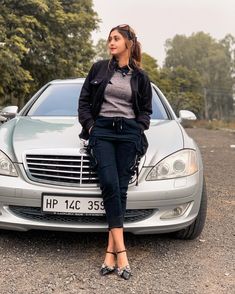 a woman sitting on the hood of a silver car in front of trees and bushes