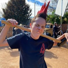 a young man holding a baseball bat with red mohawks on his head and wearing a black t - shirt