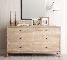 a dresser with a mirror, lamp and pictures on it next to a window in a white room