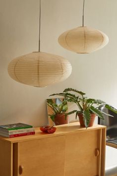 two lamps hanging from the ceiling above a wooden cabinet with books on it and plants in pots below