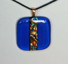 a blue square pendant with an orange and green design hanging from a black cord on a white surface