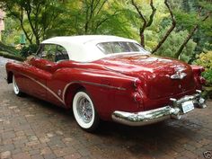 an old red and white car parked in front of some trees on a brick road