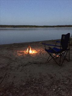 a campfire is lit up on the shore of a lake at night with chairs around it