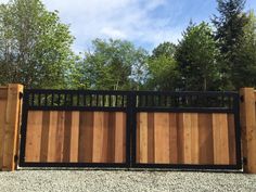 a wooden gate with black iron railings in front of some trees and gravel on the ground