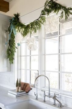 a kitchen sink under a window covered in greenery and snowflake garlands