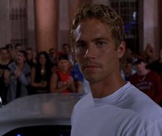 a man in white shirt standing next to a car with people looking on from behind it