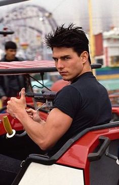 a man sitting on top of a red car in front of a ferris wheel at an amusement park