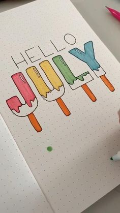 someone is writing hello july on a notebook with crayons and pencils next to it