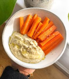 a white bowl filled with carrots and hummus next to a potted plant