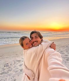 a man and woman wrapped up in blankets on the beach at sunset, taking a selfie