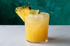 a pineapple and lime drink in a glass on a white table with a green wall behind it