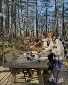 two women are sitting at a picnic table in the woods, one is serving food to the other