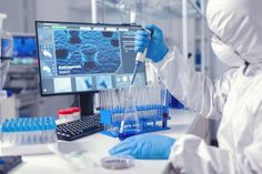 a man in white lab coat and blue gloves working on a computer screen with test tubes