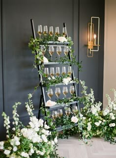 a wine rack filled with glasses and greenery on top of a wooden floor next to white flowers