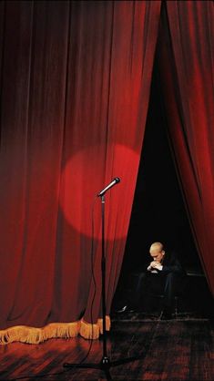 a man sitting in front of a red curtain next to a microphone on top of a wooden floor