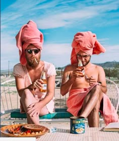 two men in towels sitting on the beach eating pizza and drinking beer