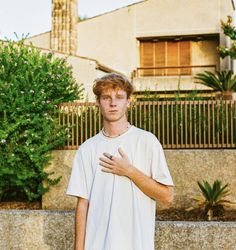 a young man standing in front of a building with his hand on his chest and looking at the camera