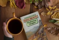 a person holding a cup of coffee next to a book on a table with autumn leaves