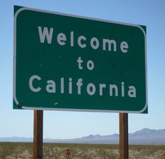 a green welcome to california sign in the middle of an open desert area with mountains in the background