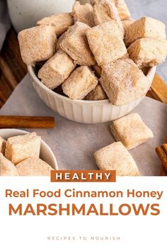 A photo of a large bowl overflowing with several cinnamon dusted 1 inch square homemade marshmallows, with a smaller bowl of marshmallows in front of it, cinnamon sticks sitting on top of parchment paper on a wooden cutting board and a ceramic jar in the background, with text that says Healthy Real Food Cinnamon Honey Marshmallows.