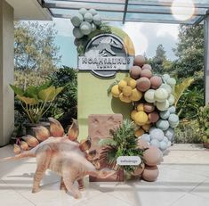 a fake dinosaur is standing in front of a sign with balloons and plants on it