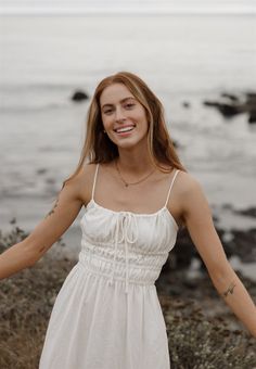 a woman in a white dress is standing by the water with her arms spread out