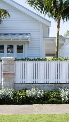 a white picket fence in front of a house with palm trees and flowers around it