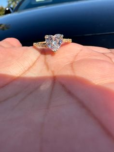 a person's hand holding a ring with a heart shaped diamond on it in front of a car