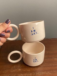 a woman's hand holding two coffee mugs on top of a wooden table