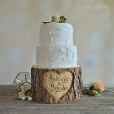 a white wedding cake on top of a tree stump with a heart shaped wooden sign