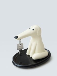 a white dog figurine sitting on top of a black plate with a key in it's mouth