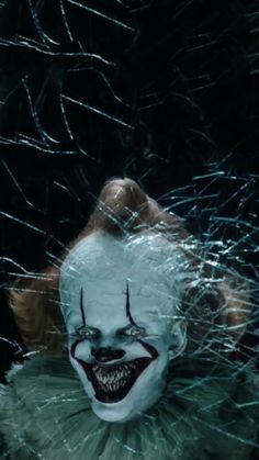 a creepy clown with white makeup and green hair is looking through the shattered glass in front of him