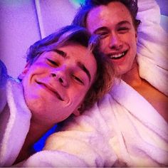 two young men laying in bed wrapped in white towels, one is smiling and the other has his eyes closed