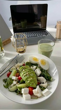 a white plate topped with salad next to a glass of green liquid and a laptop