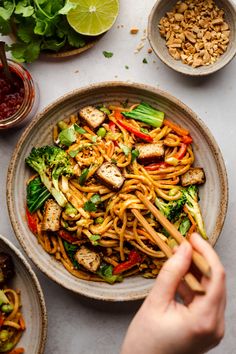 someone is holding chopsticks over a plate of noodles with vegetables and tofu