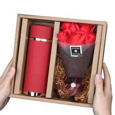 two hands holding open a red flask bottle in a cardboard box with flowers inside