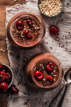 two bowls filled with chocolate pudding and cherries on top of a wooden table next to spoons