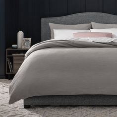 a bed with grey linens and pillows in a dark colored room, next to a night stand