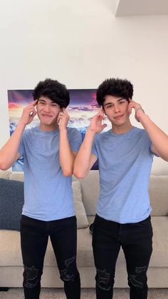 two young men standing next to each other in front of a couch with their hands on their ears