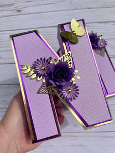 a hand holding two purple and gold cards with flowers on them in front of a white wooden background