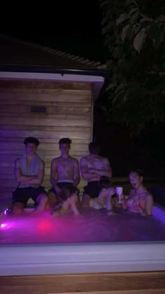 four people sitting in a hot tub at night with purple lights on the side and one person standing next to them