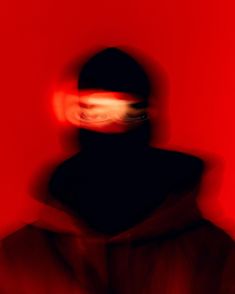 a blurry image of a person wearing a black hat and hoodie in front of a red wall