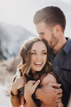 a man holding a woman in his arms and smiling at the camera with mountains in the background