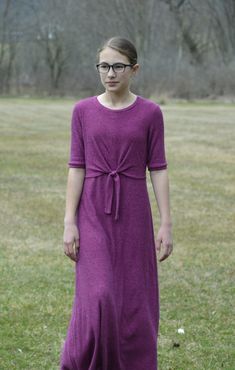 a woman standing in the grass wearing glasses and a purple dress with a tie around her waist